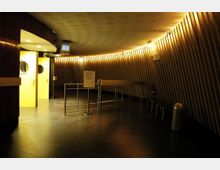 Runder Eingangsbereich in einem modernen Gebäude mit wellenförmigen Holzwänden, gelb beleuchteten Türen mit runden Fenstern, schwarzem Fliesenboden und einer Absperrung aus Metall. Warmes Licht strahlt von der Decke und den Wänden.