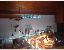 Wohnraum mit Holzdecke und großem Fenster mit buntem Glasstreifen. Der Raum ist möbliert mit Holzsesseln, Regalen und Dekorationsgegenständen, darunter Statuen und Bilderrahmen, sowie moderner Hängelampenbeleuchtung.