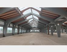 Weitläufige, leere Lagerhalle mit einem Dach aus Glaspaneelen, das auf großen, grauen Betonbögen ruht. Die Halle verfügt über seitliche Fensterfronten und eine sichtbare Holzunterkonstruktion im Dach.