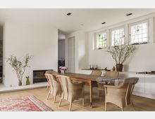 Esszimmer in einem modernen Wohnbereich mit weiß gestrichenen Wänden, großen Fenstern mit Sprossenelementen und einem Holztisch, umgeben von Korbstühlen. Im Hintergrund befinden sich ein Kamin, Pflanzen in Vasen sowie ein Teppich auf einem Holzboden.