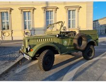 Ein olivgrüner Oldtimer-Geländewagen mit Ersatzrad an der Seite steht auf einer gepflasterten Fläche vor einer historischen, beigefarbenen Fassade mit großen Fenstern und dekorativen Elementen. Im Hintergrund ist ein weiteres monumentales Gebäude zu sehen.