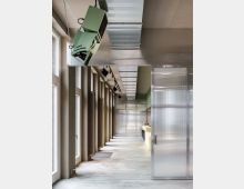 Moderner Innenraum mit hohen Fenstern auf der linken Seite, einer glatten Betonbodenfläche und sichtbaren technischen Elementen an der Decke, darunter ein grünes Gerät. Rechts befinden sich halbtransparente Trennwände aus Kunststoff.