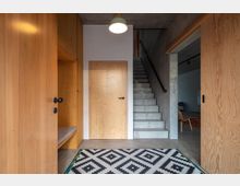 Flur in einem modernen Innenraum mit Betonwänden, Holzverkleidung und einer Treppe aus Beton. Links befindet sich ein Holzschrank mit eingebauter Sitzbank, in der Mitte eine Holztür, und der Boden ist mit einem schwarz-weiß gemusterten Teppich dekoriert.