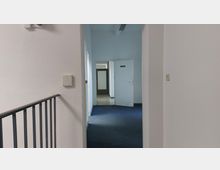 Ein weißer, schlicht gestalteter Innenflur mit Teppichboden in Blau. Am Ende des Flurs befindet sich eine halb geöffnete Tür mit einem „Ausgang“-Schild sowie eine weitere Tür aus Glas mit Sichtschutz. Links ist ein Metallgeländer sichtbar.