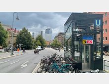 Eine Straßenszene vor einem modernen gläsernen Zugang zur U-Bahn-Station "Rosa-Luxemburg-Platz" der Linie U2, umgeben von parkenden Fahrrädern. Im Hintergrund sind Wohnhäuser, grüne Bäume und ein hoher Büroturm zu sehen, bei bewölktem Himmel.