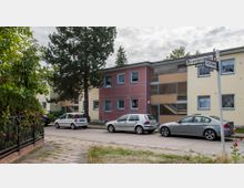 Straßenszene mit mehrstöckigen Wohngebäuden in hellgelber und rot-brauner Farbe, mit großen Fenstern und Treppenhäusern. Parkende Autos stehen am Straßenrand, links im Bild befindet sich ein Zaun mit grünem Bewuchs, und ein Straßenschild weist auf die Kreuzung von "Bospurusstraße" und "Gallipoliweg" hin.