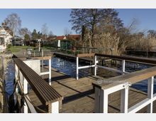 Holzsteg mit Geländer und Sitzbänken an einem kleinen Gewässer, umgeben von Schilf und Wohnhäusern im Hintergrund. Die Szene zeigt eine ruhige, ländliche Umgebung mit klarem Himmel und vereinzelten Bäumen.