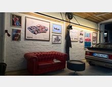 Innenraum einer Garage mit weiß verputzten Wänden und Holzbalkendecke, dekoriert mit gerahmten Auto-Postern und technischen Zeichnungen. Vor der Wand steht ein rotes Ledersofa mit kleinem Beistelltisch, daneben ist das Heck eines geparkten Autos zu sehen.