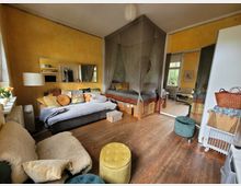 Wohnzimmer mit Holzboden, gelb gestrichenen Wänden und einem gemütlichen Sitzbereich, bestehend aus einem grauen Sofa mit bunten Kissen und einem Sessel mit Schaffell. Im Hintergrund befindet sich ein Betthimmel über einer erhöhten Liegefläche, daneben ein großer Spiegel, der den Raum optisch vergrößert.