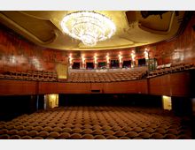 Innenansicht eines historischen Theaters mit einer Bühne im Hintergrund, umgeben von Reihen gepolsterter Stühle. Die Decke ist mit einem großen Kronleuchter geschmückt, und die Wände sind mit dekorativen Malereien verziert.