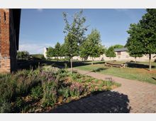 Ein gepflegter Gartenbereich mit blühenden Pflanzen im Vordergrund, einem gepflasterten Weg und mehreren jungen Bäumen. Im Hintergrund befinden sich eine Rasenfläche mit Picknick-Tischen und ein Gebäudekomplex aus Ziegel- und Putzfassaden.