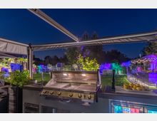 Ein beleuchteter moderner Außenbereich bei Nacht mit einem Grill im Vordergrund, umgeben von Pflanzen und einer überdachten Terrasse. Im Hintergrund sind ein Garten mit bunten Lichtern, Palmen, eine Bar und Sitzmöglichkeiten zu sehen.