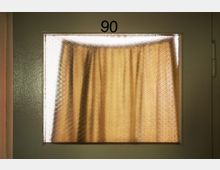 Eine beigefarbene Tür mit einem kleinen rechteckigen Milchglasfenster, hinter dem ein gelber Vorhang sichtbar ist. Über dem Fenster steht die Zahl "90".