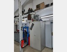 Innenraum einer Werkstatt mit weiß gestrichenen Wänden und abgerundeter Decke. An der Wand befinden sich Regale mit Werkzeugen, alten Gegenständen und Schildern. Ein großer Metallschrank, eine hydraulische Presse in Blau, eine Werkbank mit Schubladen sowie ein Feuerlöscher sind sichtbar.