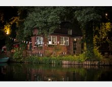 Backsteingebäude an einem Flussufer, umgeben von Bäumen und dekoriert mit Wimpelketten. Im Vordergrund spiegelt sich das Gebäude im Wasser, während Lichter die Szene dezent beleuchten.