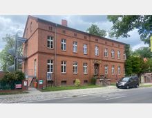 Backsteingebäude mit klassischer zweigeschossiger Architektur, mehreren rechteckigen Fenstern und einer Außentreppe auf der rechten Seite. Vor dem Gebäude befindet sich eine kleine Grünfläche, ein Gehweg und eine Straße, daneben ein geparktes Auto und eine Bushaltestelle.