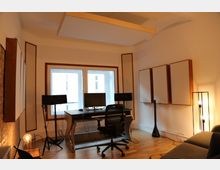 Ein modernes Arbeitszimmer mit einem Schreibtisch, zwei Monitoren, einem MIDI-Keyboard und Studiolautsprechern vor einem großen Fenster. Akustikelemente an den Wänden, eine Hängedecke zur Schallabsorption, eine Stehlampe und ein Sofa ergänzen die Einrichtung, während der Holzboden den Raum wärmt.
