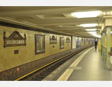 U-Bahn-Station "Fehrbelliner Platz" mit gelb gekachelten Wänden und reich verzierten Stationsschildern; seitliche Ansicht eines geschwungenen Bahnsteigs mit Gleisen und einem einzelnen Fußgänger.