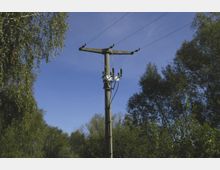 Strommast mit Oberleitungen in einer ländlichen Umgebung, umgeben von Bäumen und Sträuchern, unter einem klaren blauen Himmel.