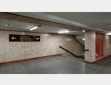Ein unterirdischer Bahnhofsgang mit beigefarbenen Wandfliesen, Deckenbeleuchtung und grauem Fliesenboden. Eine Treppe mit schwarzem Handlauf führt nach unten, daneben befindet sich ein orangefarbenes Gitter; ein Schild mit Richtungshinweisen ist oberhalb angebracht.