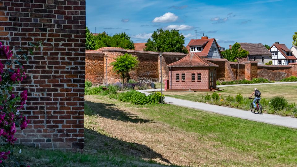 Grünanlage mit gepflastertem Weg vor einer langen, historischen Backsteinmauer und einem kleinen Backsteingebäude mit Walmdach. Im Hintergrund stehen Fachwerkhäuser und Bäume; links ist eine Backsteinwand mit blühenden Sträuchern zu sehen.