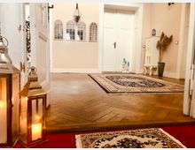 Ein stilvoll eingerichteter Flur mit hellen Wänden und einem hölzernen Fischgrätboden. Der Raum enthält dekorative Teppiche, eine weiße doppelte Holztür, goldene Laternen mit Kerzen, sowie ein kleines weißes Regal mit Trockenblumen und Kerzenständern.