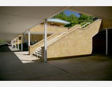 Überdachter Durchgang mit gelben Kachelmauern und einer breiten Außentreppe, die zu einer oberen Ebene führt. Im Hintergrund sind Gebäude mit Flachdächern sowie grüne Bäume sichtbar.