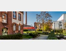 Backstein-Gebäude im Vordergrund mit hellen Fenstereinfassungen, umgeben von gepflasterten Wegen, grünen Büschen und Bäumen; im Hintergrund ein roter Zug, Gleise und ein hoher Schornstein im Industriegebiet.