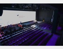 Großer moderner Veranstaltungsraum mit einer Bühne, LED-Hintergrundwand und mehreren Mikrofonen und Instrumenten darauf, umgeben von Reihen mit schwarzen Stühlen. Der Raum ist dunkel mit violetter Beleuchtung auf dem Boden und wird für eine Präsentation oder Aufführung vorbereitet.