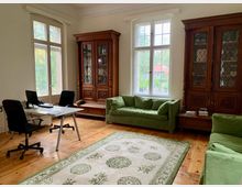 Ein heller Raum mit Holzboden, zwei großen Fenstern und Blick ins Grüne. Der Raum ist eingerichtet mit zwei großen Holzvitrinen, einem weißen Schreibtisch mit zwei schwarzen Bürostühlen sowie grünen Polstersofas und einem cremefarbenen Teppich mit grünem Muster.