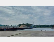 Industriehafen mit einem befestigten Kai, an dem ein Wasserweg verläuft. Im Hintergrund sind Lagergebäude, ein großer Holzspänhaufen, ein Schornstein und bewaldete Flächen sichtbar; rechts am Bildrand liegt ein kleines Schiff am Ufer.