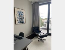 Modernes Büro oder Wohnraum mit einer schwarzen Ledersessel-Liege auf einem hellen, runden Teppich, einem großen Fenster mit grauen Vorhängen und Blick auf einen Balkon. An der Wand hängt ein abstraktes Kunstwerk in Blau und Weiß, ein Schreibtisch mit Tastatur und Stuhl steht im Vordergrund.