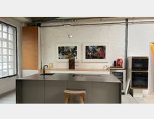 Moderne Küchenzeile in einem loftartigen Raum mit weißen Backsteinwänden, großer Sprossenfensterfront und freiliegenden Leitungen an der Decke. Zentral steht eine graue Kücheninsel mit Spüle und Kochfeld; an der Wand hängen zwei gerahmte Kunstbilder, daneben sind eingebaute Backöfen und ein kleiner Beistelltisch.