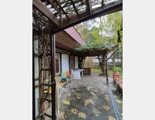 Überdachte Terrasse mit Natursteinboden, angrezendem Holzgeländer und überwuchertem Pergola-Dach, dekoriert mit Gartenmöbeln in bunten Farben. Im Hintergrund befindet sich ein kleiner Garten mit Bäumen und Sträuchern.