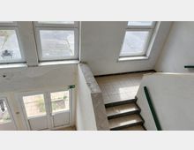 Treppenhaus mit weißen Wänden und großen, rechteckigen Fenstern. Die Treppe ist mit hellen Fliesen belegt, das Geländer ist grün, und im unteren Bereich ist eine Glastür mit einem Notausgangsschild sichtbar.