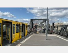 Gelbe U-Bahn mit geöffneten Türen an einem Hochbahnsteig, im Hintergrund eine Stahlbrücke mit Schienen, Signalanlagen und umliegender moderner Bebauung unter einem Himmel mit Wolken.