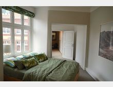 Schlafzimmer mit großem Fenster, durch das eine Ziegelsteinfassade eines Nachbargebäudes sichtbar ist. Im Raum stehen ein Bett mit grüner Bettwäsche und gemusterten Kissen, ein kleiner Nachttisch mit Lampe, sowie eine geöffnete Tür, die in einen angrenzenden Raum führt.