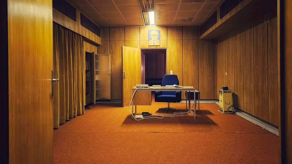 Innenraum eines retro wirkenden Büros mit holzvertäfelten Wänden und orangefarbenem Teppichboden, in der Mitte ein Schreibtisch mit Bürostuhl und sichtbaren Kabeln. Abgehängte Decke mit Leuchtstofflampe, seitlich ein Vorhang und im Hintergrund eine offene Tür sowie ein gerahmtes Porträt über der Tür.