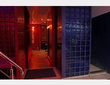 Ein schmaler, rot beleuchteter Gang mit glänzenden Glasbausteinwänden in Blau und Rot. Der Raum im Hintergrund zeigt Sitzmöbel und wird von dekorativen Lichtmustern erhellt.