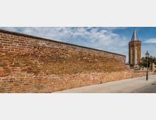 Rote Backsteinmauer mit einem geklinkerten historischen Kirchturm im Hintergrund. Der Weg davor ist gepflastert, und eine Laterne steht am rechten Bildrand unter blauem Himmel mit vereinzelten Wolken.