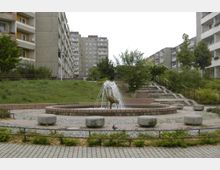 Wohngebiet mit Mehrfamilienhäusern im Hintergrund, zentral ein kleiner Brunnen mit einer Skulptur, die von Wasserstrahlen umgeben ist. Der Bereich ist von Grünflächen, Bäumen, Treppen und gepflasterten Wegen umgeben.
