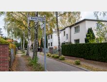 Wohnstraße mit zweigeschossigen, weißen Mehrfamilienhäusern, gesäumt von Birkenbäumen und gepflegten Hecken. Ein Bürgersteig verläuft entlang der Häuser, und ein Straßenschild mit der Aufschrift „Ruthnerweg“ ist sichtbar.