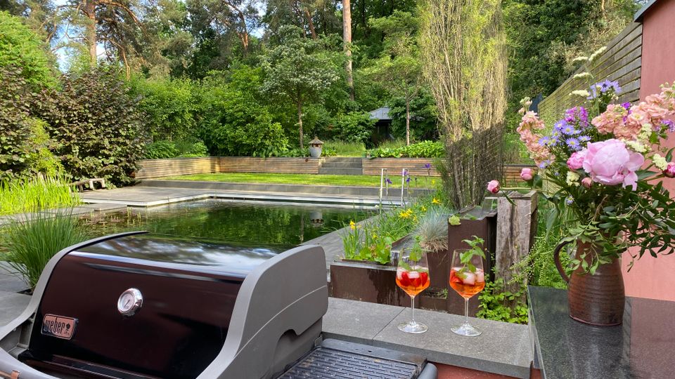 Terrasse in einem begrünten Garten mit modernem Naturpool, Holzterrassen und gestuften Beeten vor dichtem Baumbestand. Im Vordergrund stehen ein Gasgrill, zwei Getränkegläser und eine Vase mit Blumen neben einer Gartenmauer.