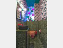 Small modern bathroom with terrazzo wall and floor tiles, a round black vessel sink with a bright red faucet, and a wall-mounted pink toilet. Patterned wallpaper and a colorful glass-block window provide vivid accents, with a mirror and globe light fixture above the vanity.
