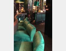 Innenansicht einer stilvollen Bar mit einer geschwungenen Bartheke und grünen Samtpolstern auf den Barhockern. Im Hintergrund sind eine beleuchtete Rückwand mit Getränkeflaschen, tropisch bedruckte Wände sowie grüne Lampen zu sehen.