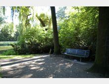 Ein schattiger Park mit Bäumen und dichtem Grün, im Vordergrund eine hölzerne Parkbank neben einem schwarzen Abfalleimer. Im Hintergrund ist ein kleiner Fluss oder Teich zu sehen.