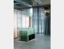 Modernes Interieur mit einem grünen Tresen aus transparenten Glasfliesen, vor einer Wand aus transluzenten Paneelen. Im Hintergrund hängen dunkle Vorhänge und eine Lautsprecherbox, ergänzt durch sichtbare Lüftungsschächte und eine Metallträgerkonstruktion an der Decke.