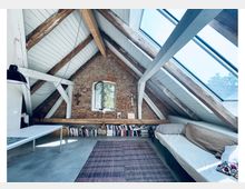 Dachgeschoss-Schlafzimmer mit schrägem, weiß verkleidetem Dach, sichtbaren Holzbalken und großen Dachfenstern, die viel Tageslicht hereinlassen. An einer Backsteinwand mit kleinem Fenster stehen hohe Bücherstapel; daneben sind Bett und Teppich in einem schmalen Raum angeordnet.