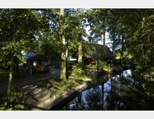 Grüne Parklandschaft mit einem kleinen Holzgebäude und einem gepflasterten Weg, der zu einer Anlegestelle an einem schmalen, von Bäumen umgebenen Wasserkanal führt. Im Hintergrund ist ein größeres Gebäude mit Reetdach sichtbar.