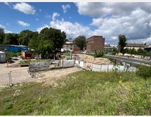 Freifläche mit Bauzäunen und einem kleinen urbanen Gartenbereich, umgeben von vereinzelten Containern und Bäumen; im Hintergrund eine Straße, ein Backsteingebäude und Graffiti-verzierte Wände. Der Himmel ist leicht bewölkt.